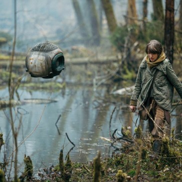 Young girl walks through a swamp accompanied by a spherical drone with a smiley face drawn on its front